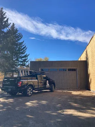 Black pickup truck parked beside a garage under a bright blue sky, with trees and shadows in the yard