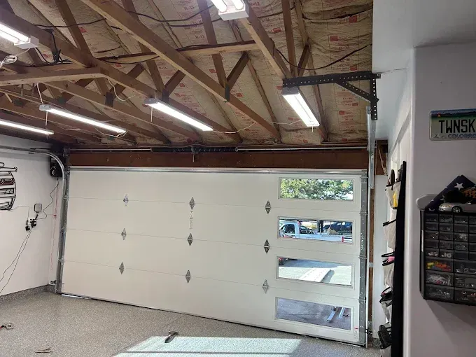 Garage interior with white overhead door, exposed rafters, fluorescent lights, and wall-mounted shelves