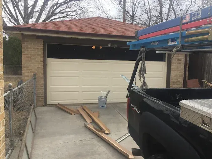 Black pickup truck loaded with lumber in a driveway in front of a garage, with wood pieces on the ground