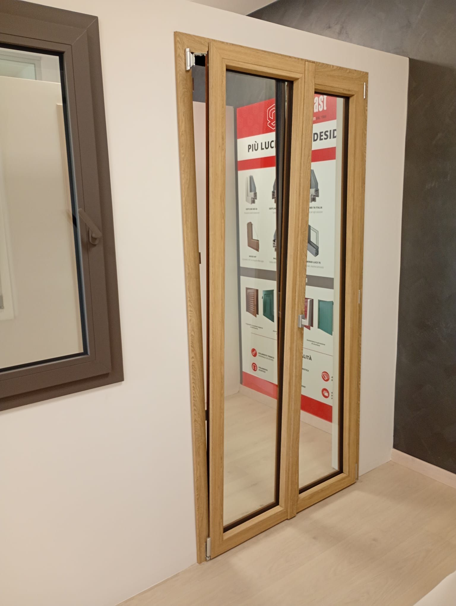 Una porta a doppia anta con venature del legno chiaro, esposta accanto a una piccola cornice di finestra in uno showroom.
