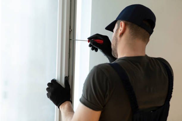 Un addetto alla manutenzione, con indosso un berretto e guanti neri, usa un cacciavite per regolare un telaio di una finestra bianca all'interno dell'abitazione.