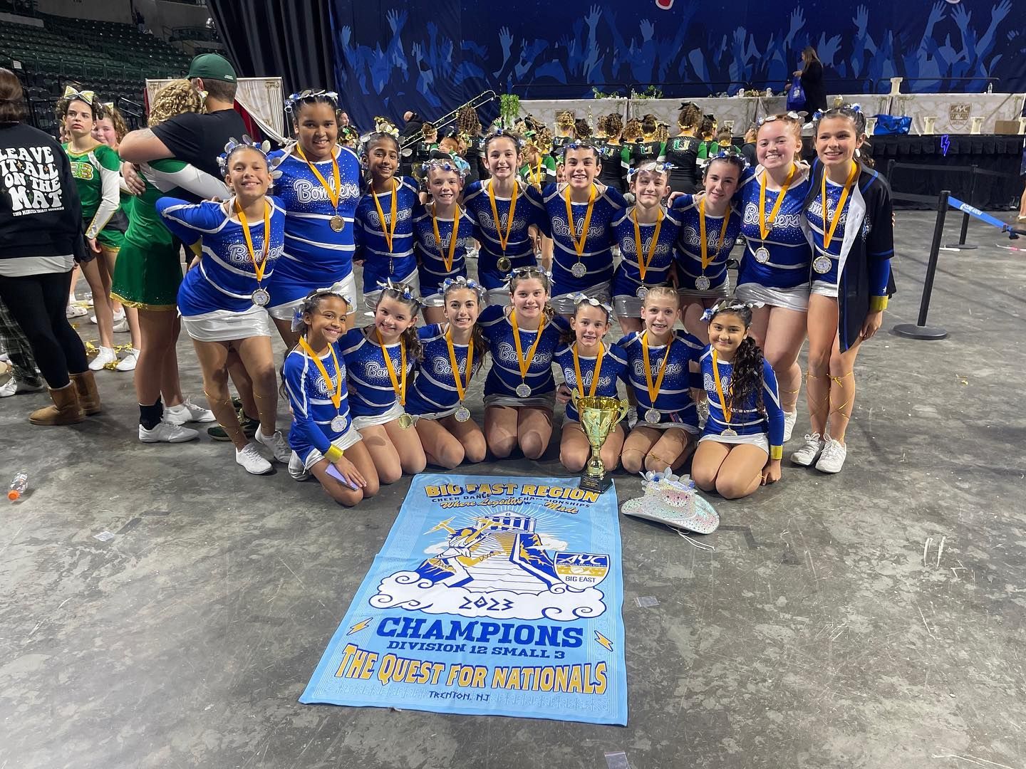 Cheerleading team in blue and gold uniforms, kneeling and standing with medals, holding championship banner.