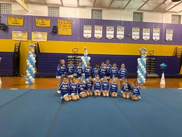 Cheerleading squad in blue and white uniforms kneels on a blue mat in a gym.