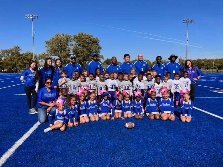 Youth football team and cheerleaders on a blue field posing for a photo under a sunny sky.