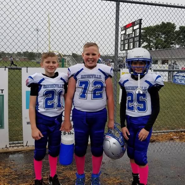 Three youth football players in blue and white uniforms, standing in front of a fence, holding gear.