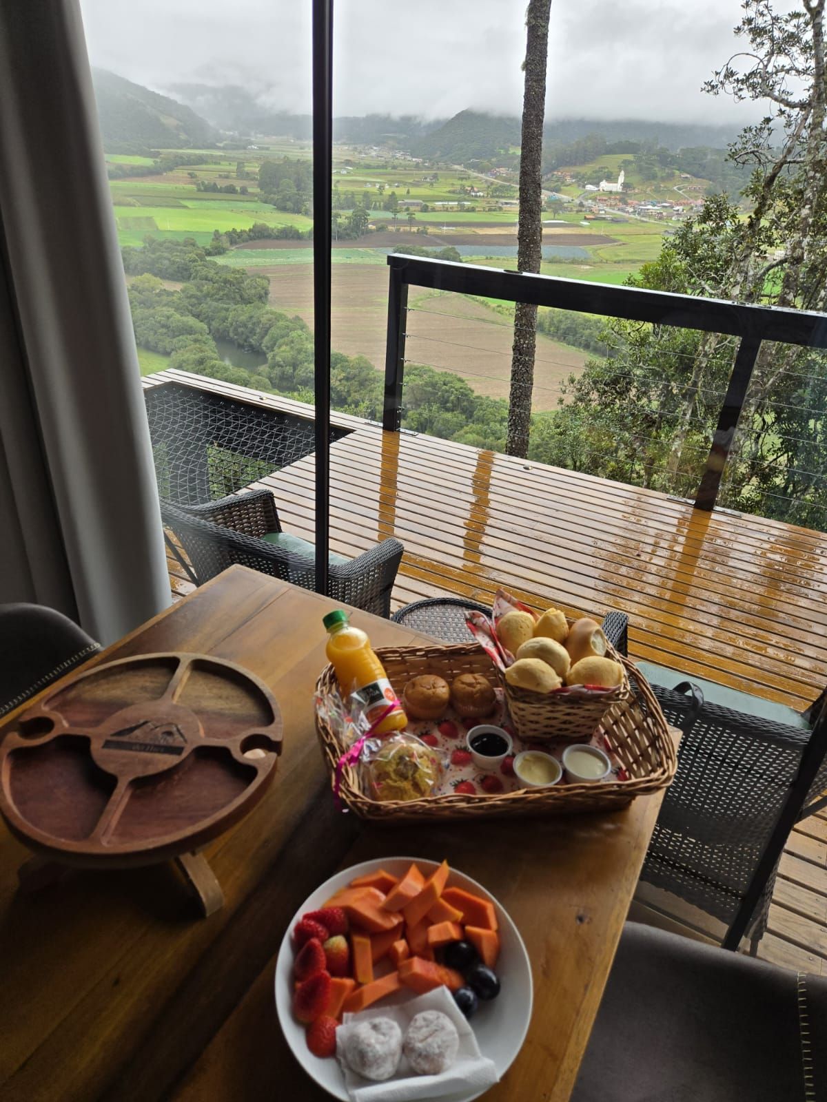 Mesa de café da manhã com vista para um vale: prato de frutas, cesta de pães, suco, tábua de servir de madeira.