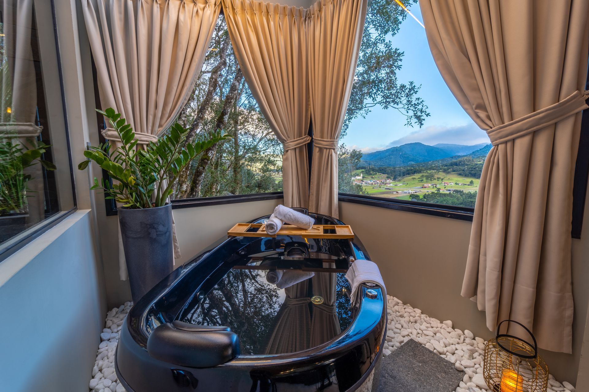 Banheira preta cheia de água, ao lado de uma janela com vista para a montanha e cortinas cor creme.