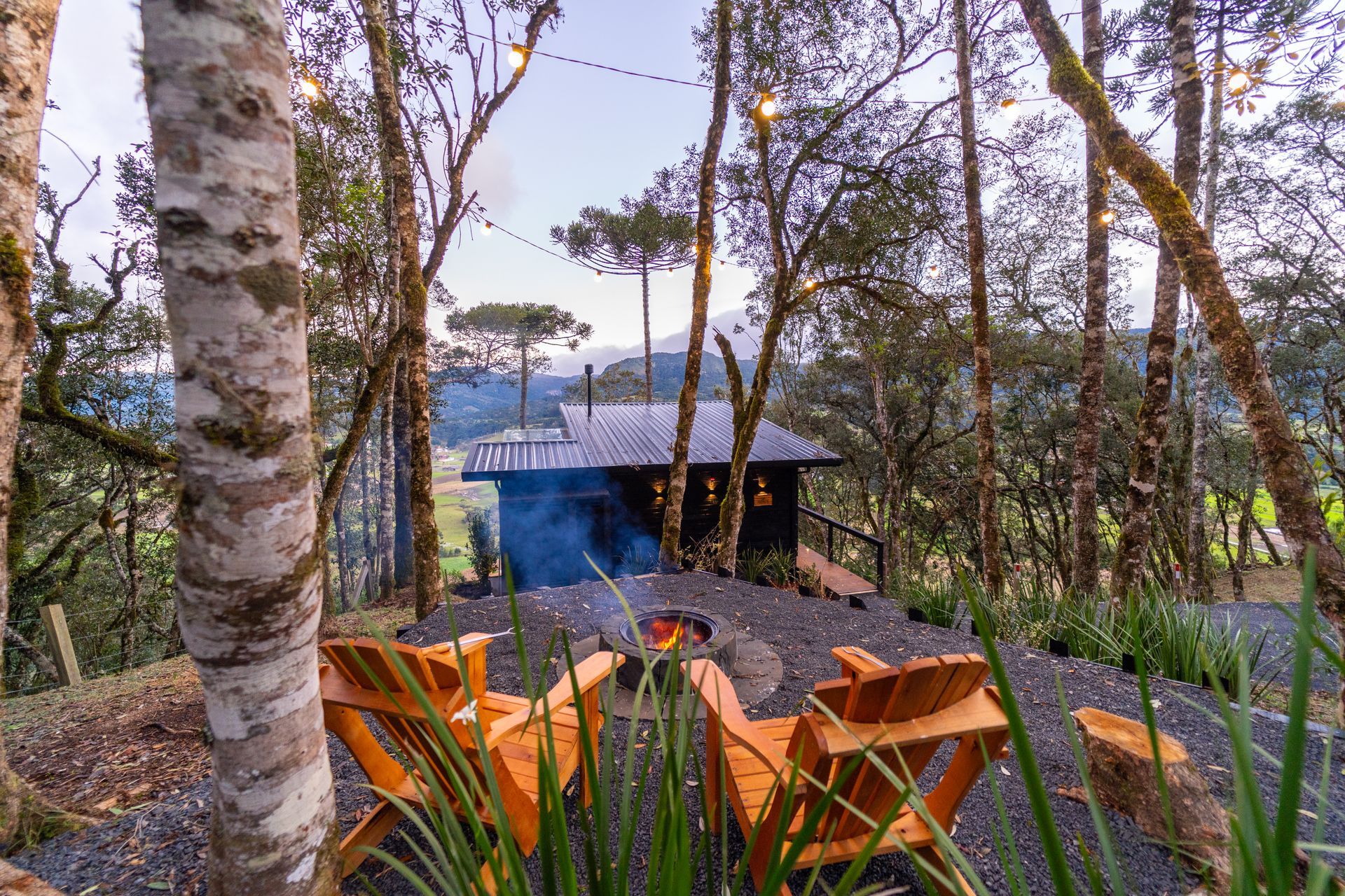 Duas cadeiras de madeira estão de frente para uma cabana com uma fogueira, cercada por árvores.