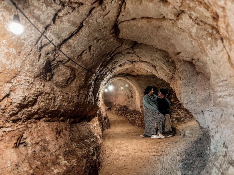 Duas figuras abraçadas dentro de uma caverna rústica em forma de arco, iluminada por luzes de corda.