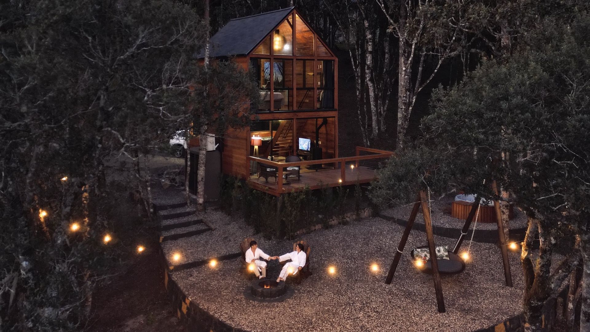 Aconchegante casa na árvore à noite, com um deck. Duas pessoas sentam-se perto de uma fogueira, rodeadas por luzes de corda.