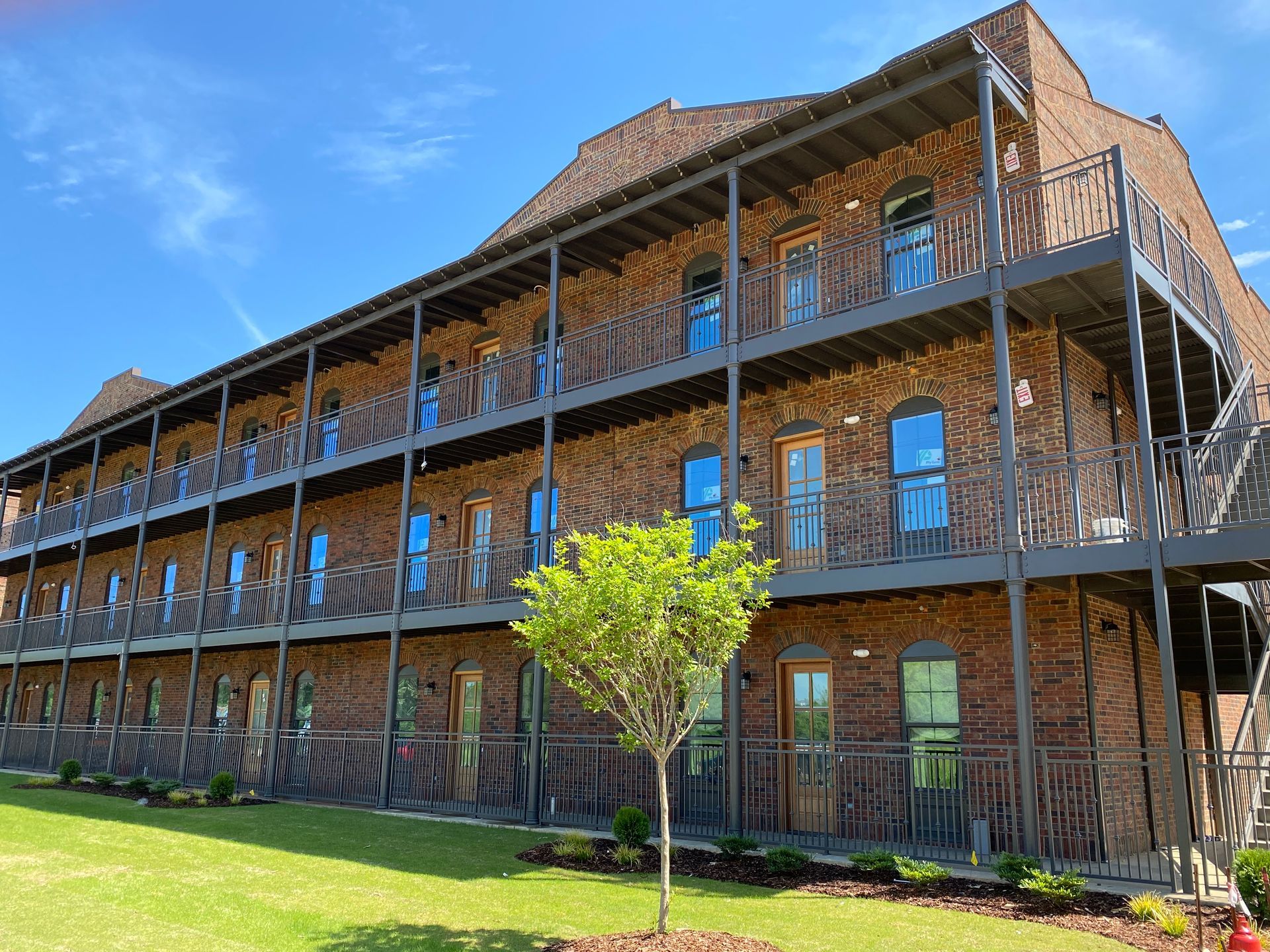 Brick building with three stories, each with a balcony. Green lawn and a small tree in front.