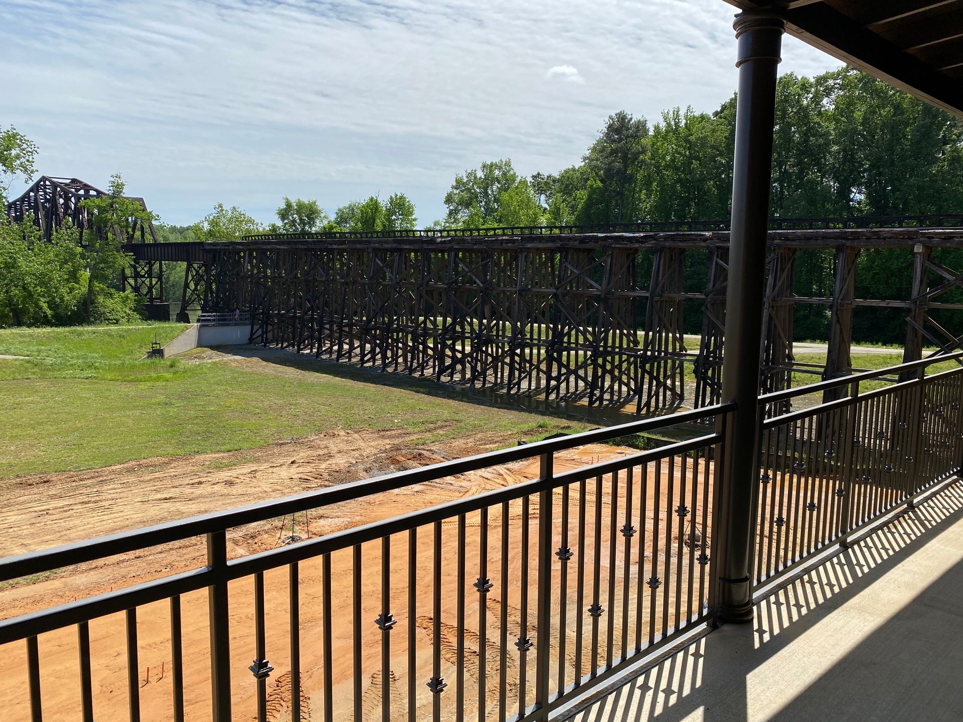 Wooden trestle bridge over a sandy area, viewed from a porch with black metal railings. Trees and blue sky in background.