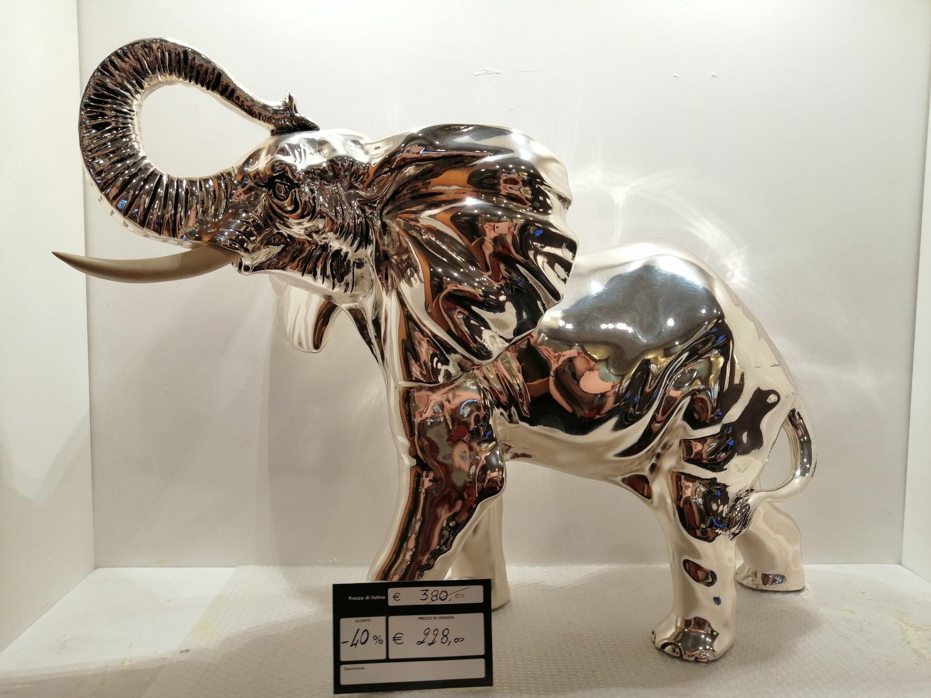 Scultura di elefante in argento, con la proboscide sollevata, appoggiata su una superficie bianca e con una piccola etichetta sul davanti.