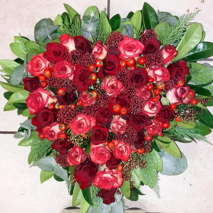 Bouquet a forma di cuore composto da rose rosse e fiori bordeaux con foglie verdi su sfondo chiaro.