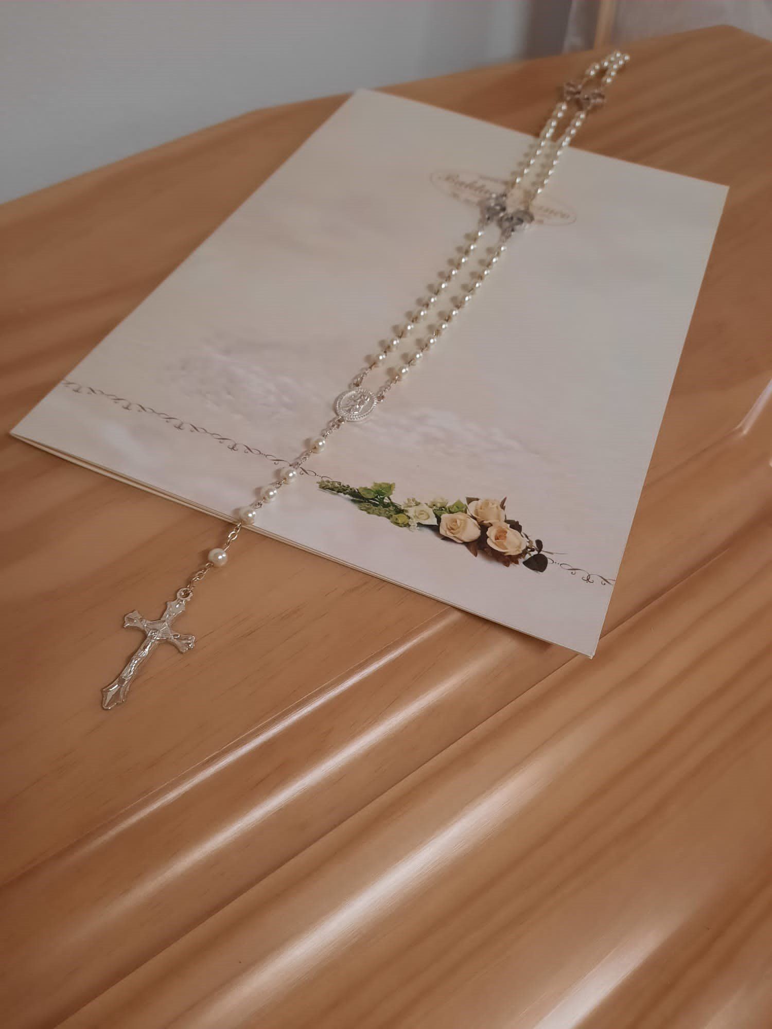 Collana del rosario adagiata in diagonale su un cartoncino bianco, sopra una superficie di legno color cuoio.