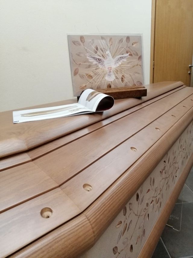 Bara di legno con un libro aperto e un biglietto floreale appoggiati sopra, in un contesto funebre.