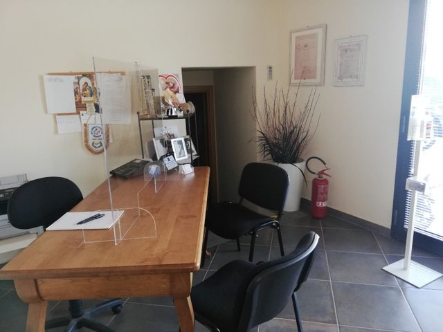Piccolo ufficio con scrivania in legno, sedie nere, scaffali e un estintore vicino alla porta.