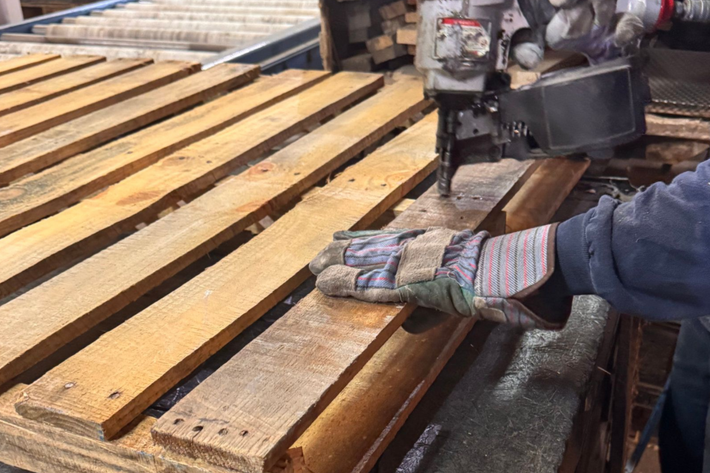 Gloved worker uses nail gun on wooden planks on a lumber conveyor in a workshop