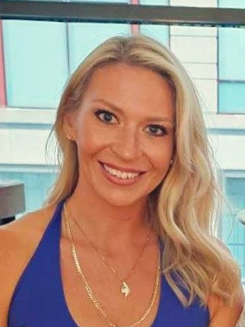 Smiling blonde woman in a blue halter top with gold necklaces, standing indoors near a window.