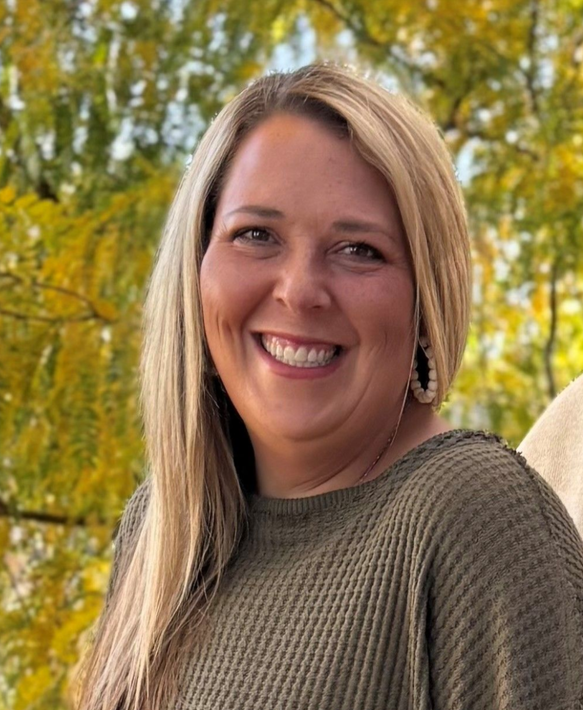 Smiling woman with blonde hair in a brown sweater, outdoors with autumn trees in the background