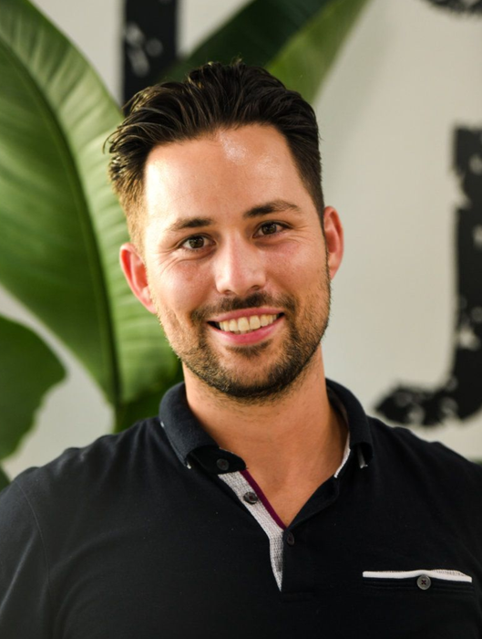 Smiling man in a black polo shirt standing indoors with green plants in the background