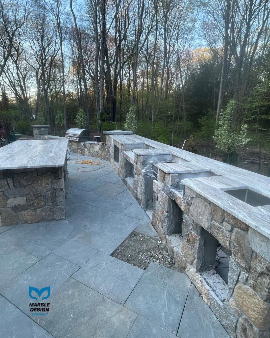 Stone patio with benches and a built-in grill area beside a wooded park path