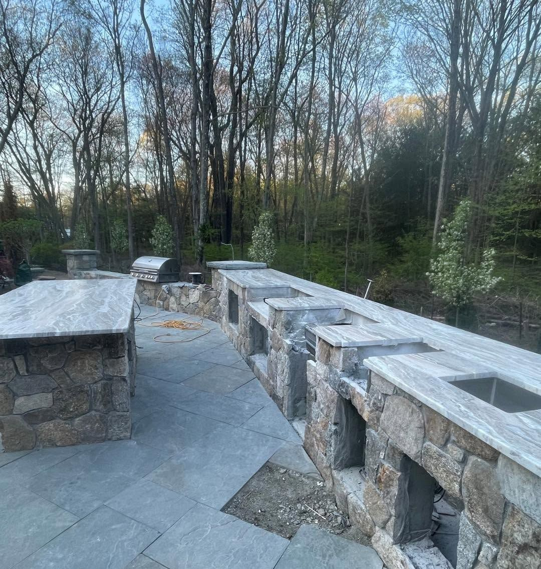 Stone patio with built-in outdoor fireplace and seating walls, beside a wooded area at dusk