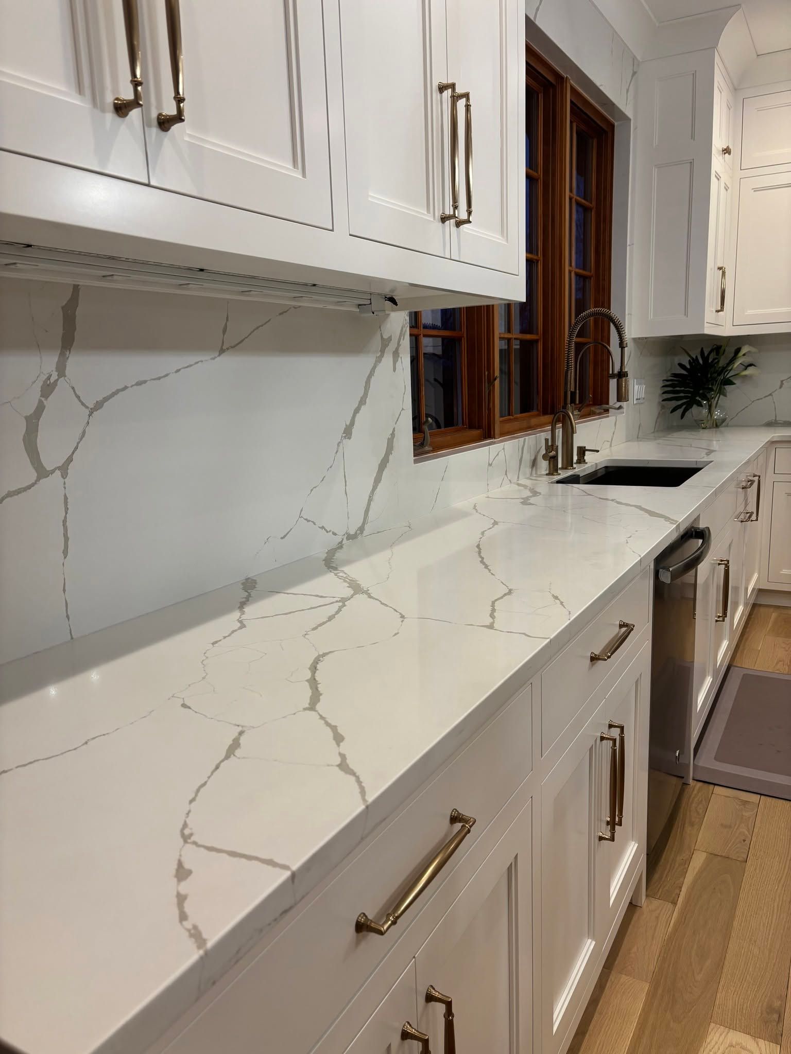 Modern kitchen counter with white marble-look countertop, white cabinets, brass handles, and a window above the sink.