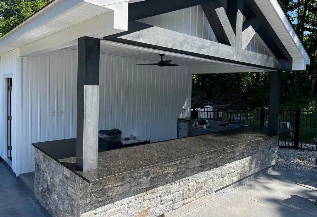 Covered stone patio with white siding, exposed beams, and a ceiling fan beside a gated yard.