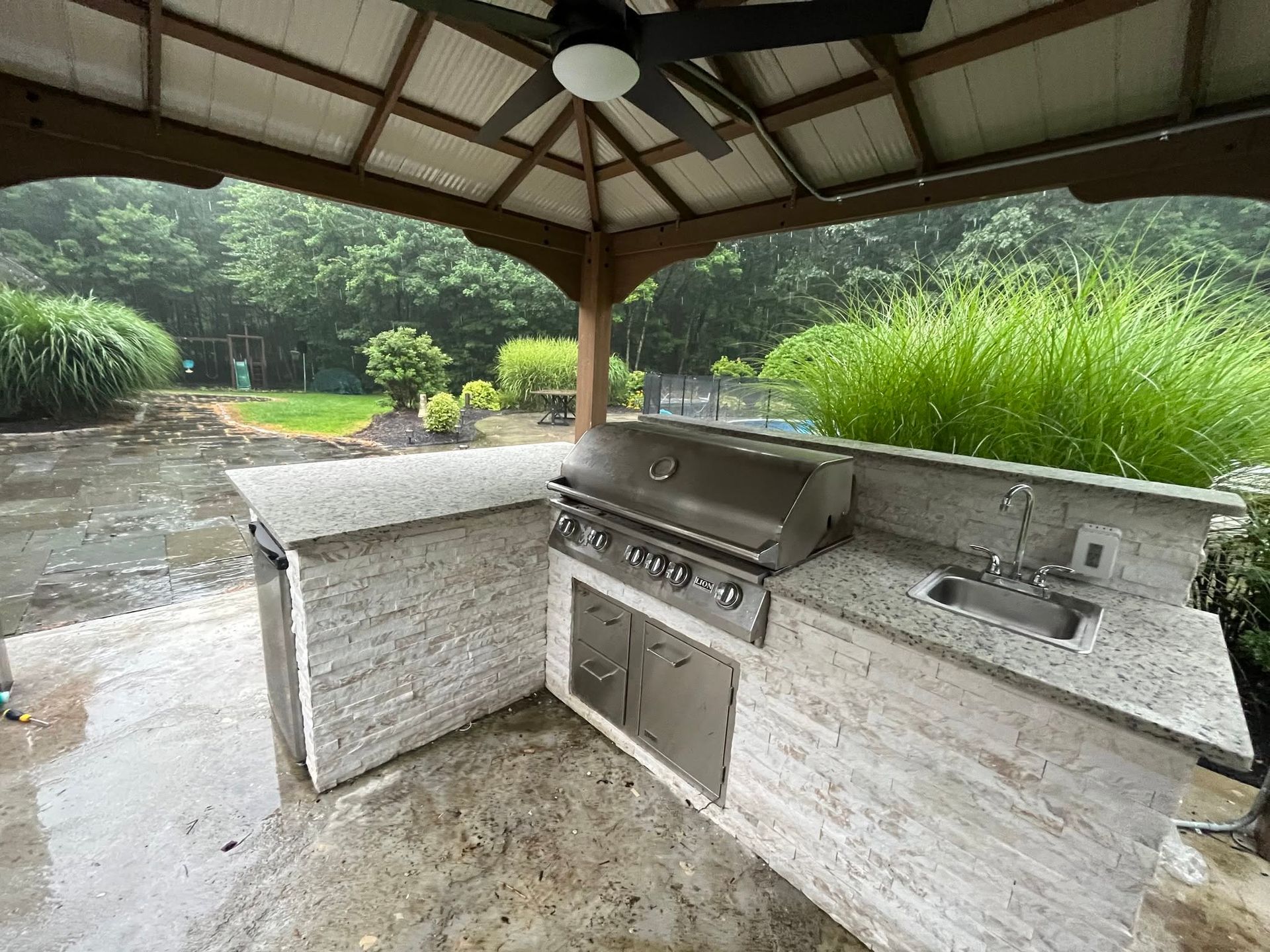 Outdoor stone grill and sink under a gazebo beside a pond and tall grasses.