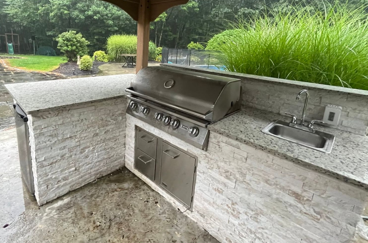 Outdoor built-in stone grill and sink under a covered patio, with greenery in the background