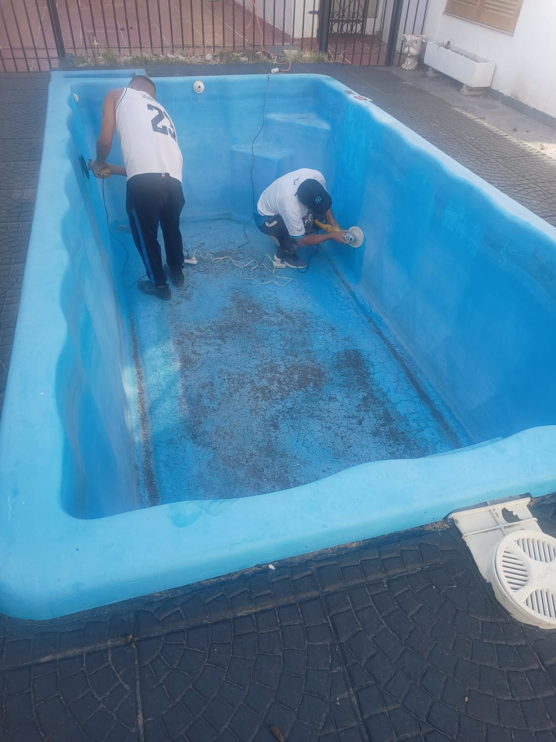 Dos hombres limpiando una piscina azul vaciada; uno de pie, otro arrodillado; al aire libre.