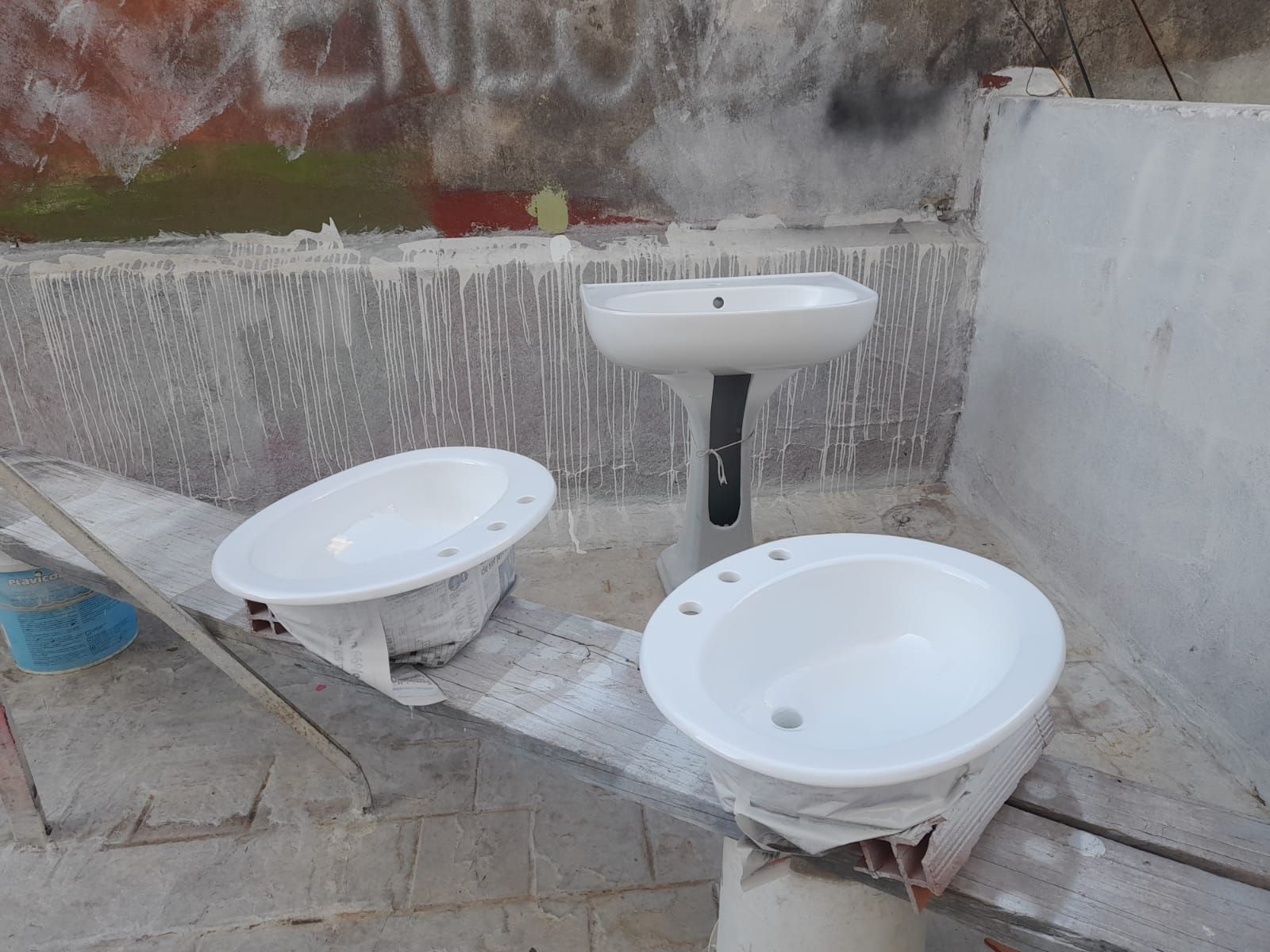 Tres lavabos de baño blancos sobre una superficie de hormigón, un lavabo de pedestal y dos lavabos de cuenco, con una pared pintada de fondo.