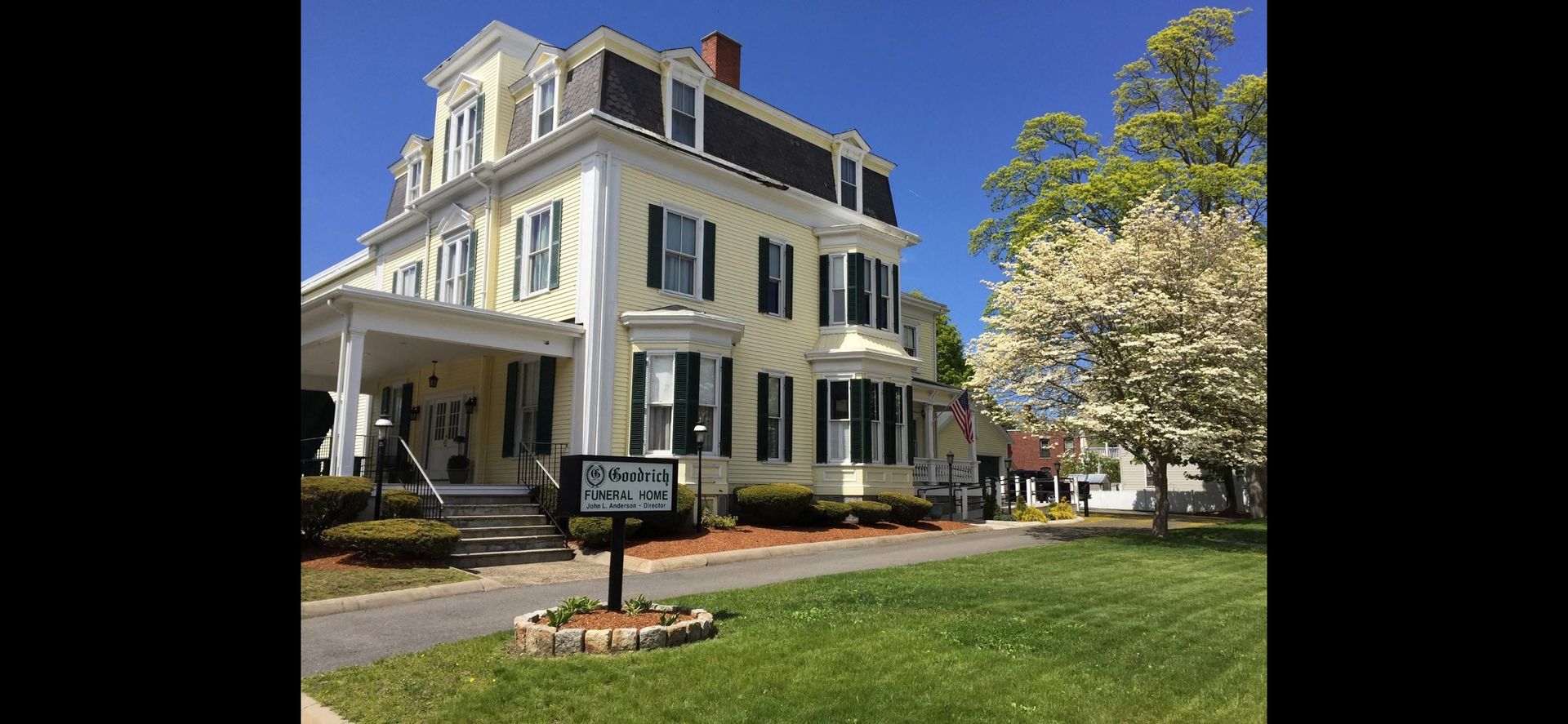 Yellow building with black roof, green shutters, porch, sign reading 