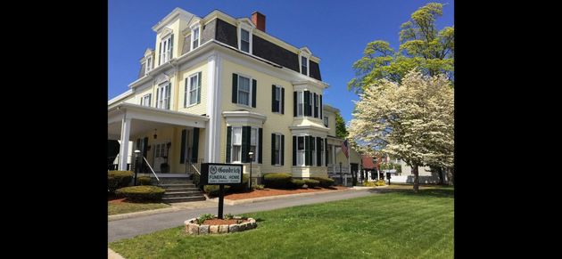 Yellow building with black roof, green shutters, porch, sign reading 