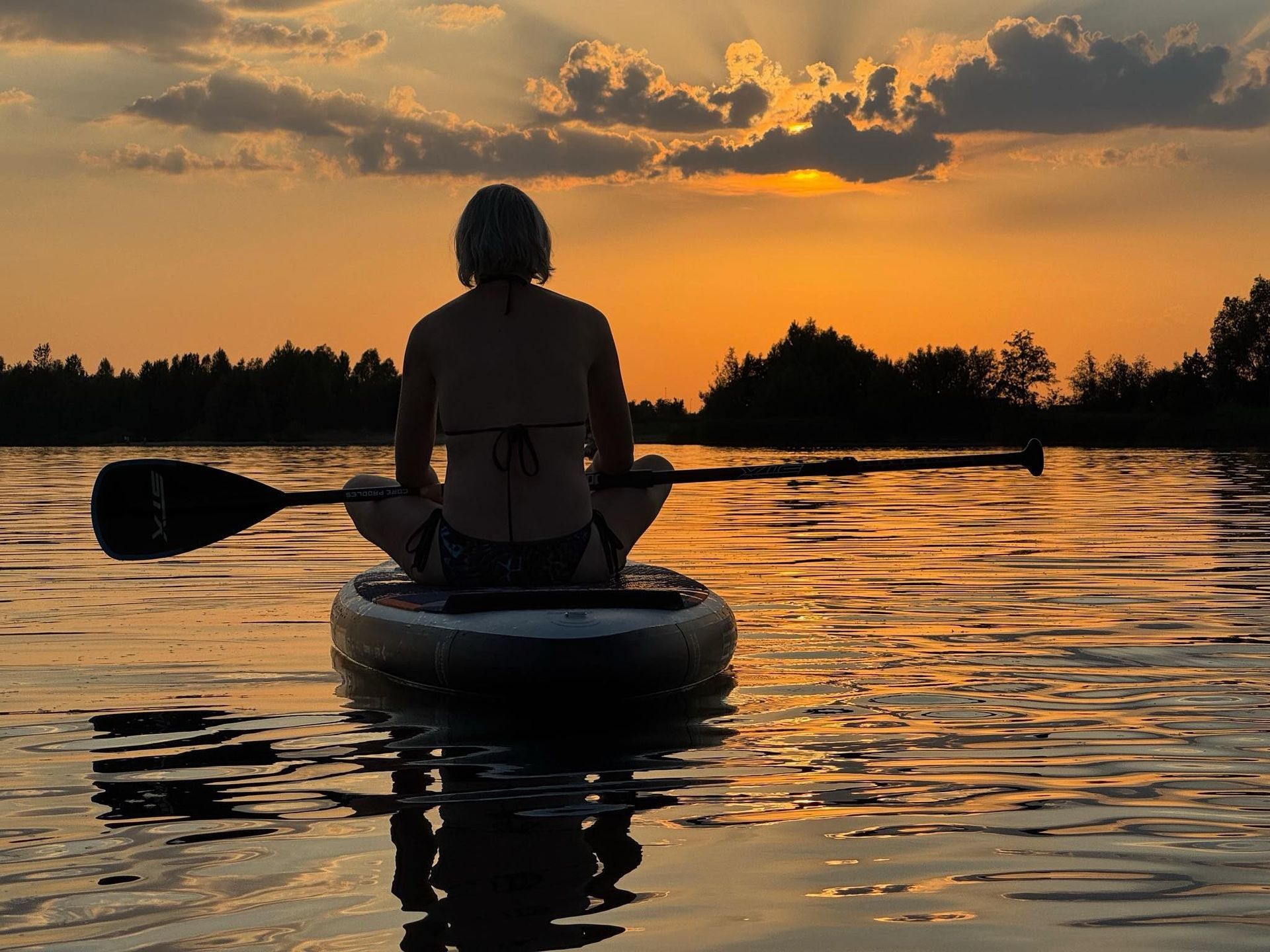 Persoon op een paddleboard bij zonsondergang, silhouet tegen een gouden hemel, peddel vasthoudend, kalm water.