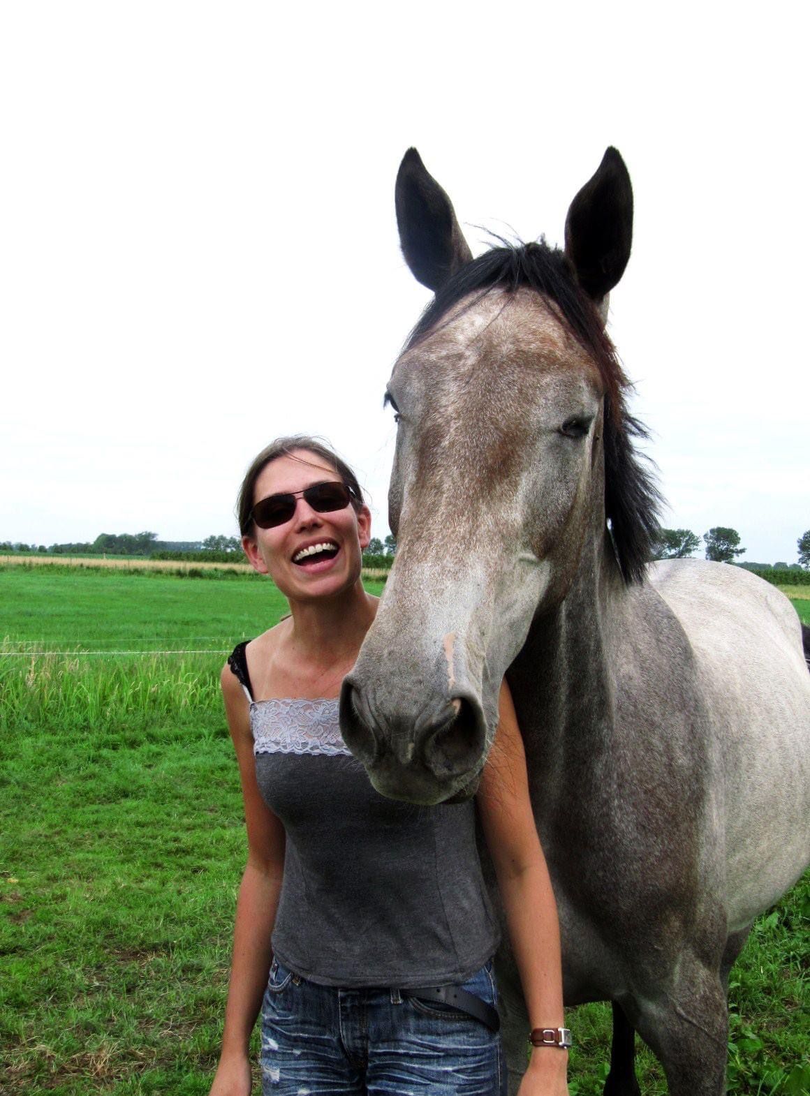 Vrouw lacht naast een grijs paard in een groen veld.