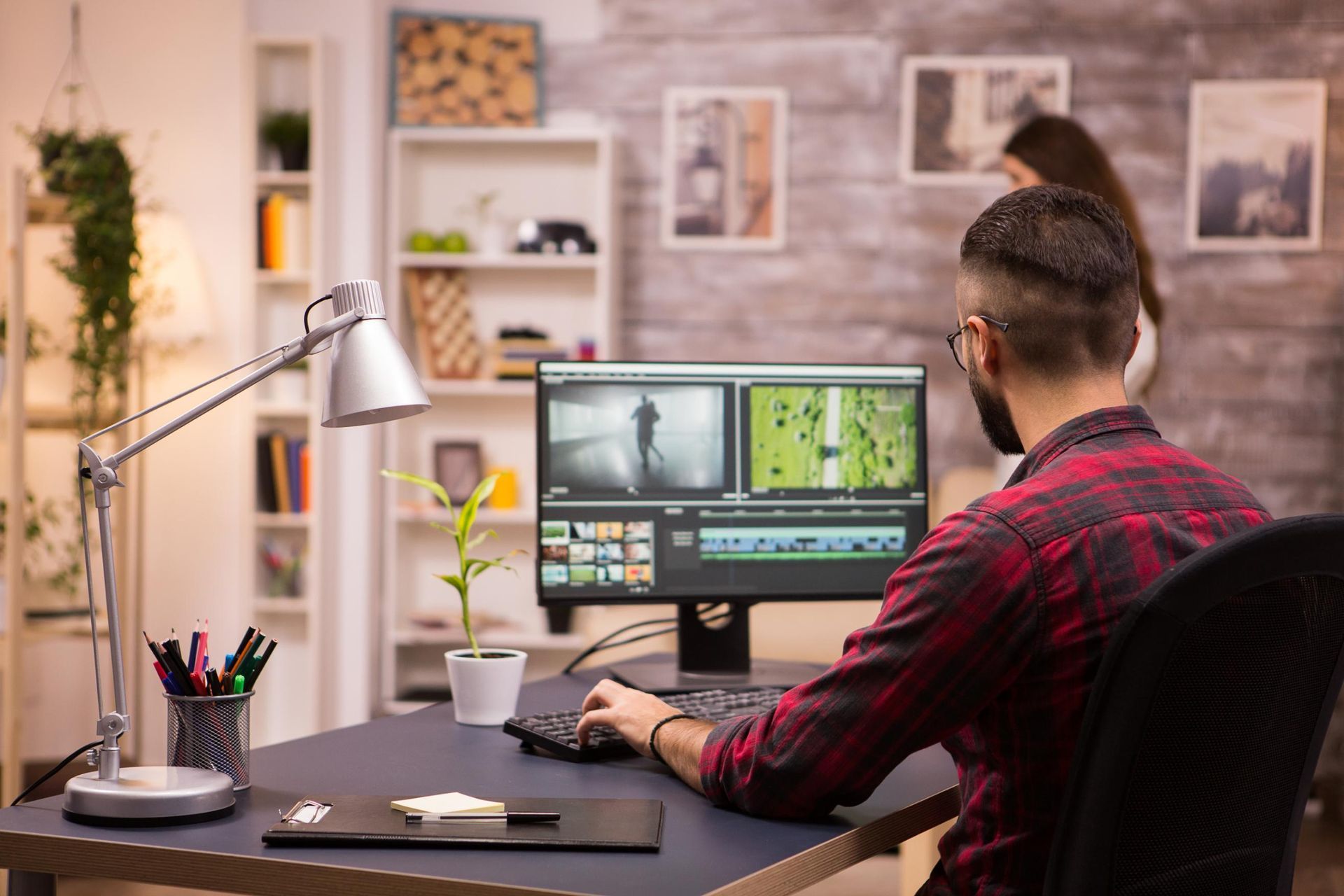 Man at desk editing video on a computer; woman stands in the background.