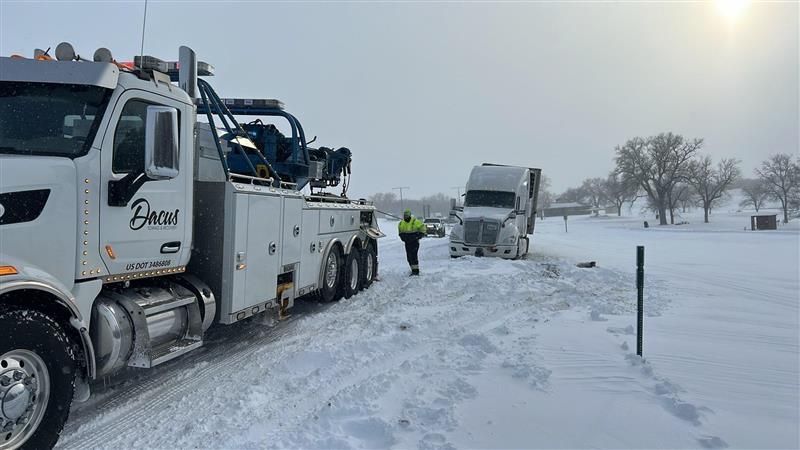 Tow truck and semi stranded on a snowy road, with a worker in a reflective jacket nearby.