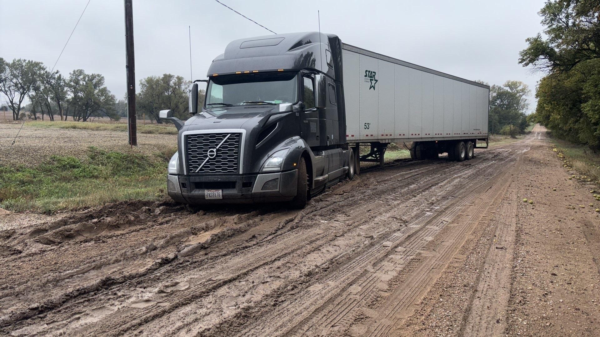 Black semi-truck with white trailer parked on a muddy dirt road in a rural area