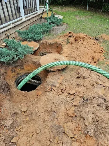 A green hose is inserted into an open, excavated septic tank access hole near a house foundation and landscaping.