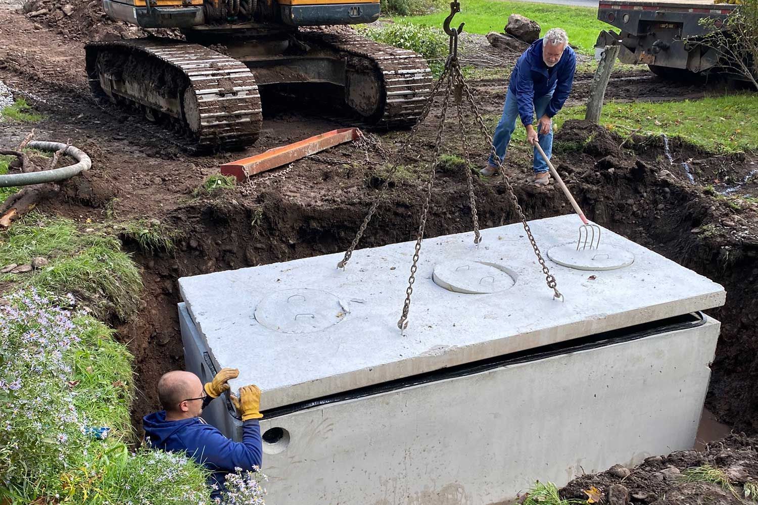 Workers install a large concrete septic tank in an excavated trench outdoors, using an excavator to lift the lid.