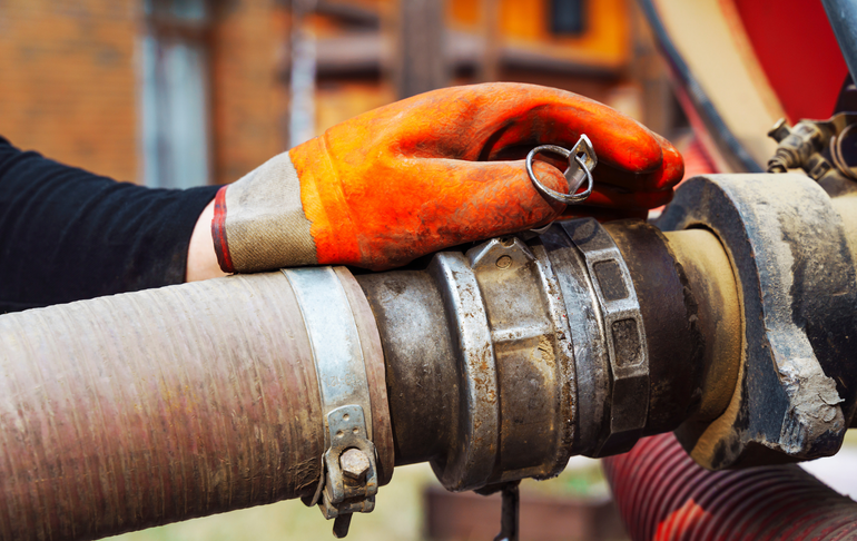 A person wearing an orange work glove holds onto a metal industrial pipe connection with a clamp.