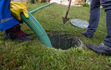 Two people use a green suction hose to pump out an open residential septic tank in a grassy yard.