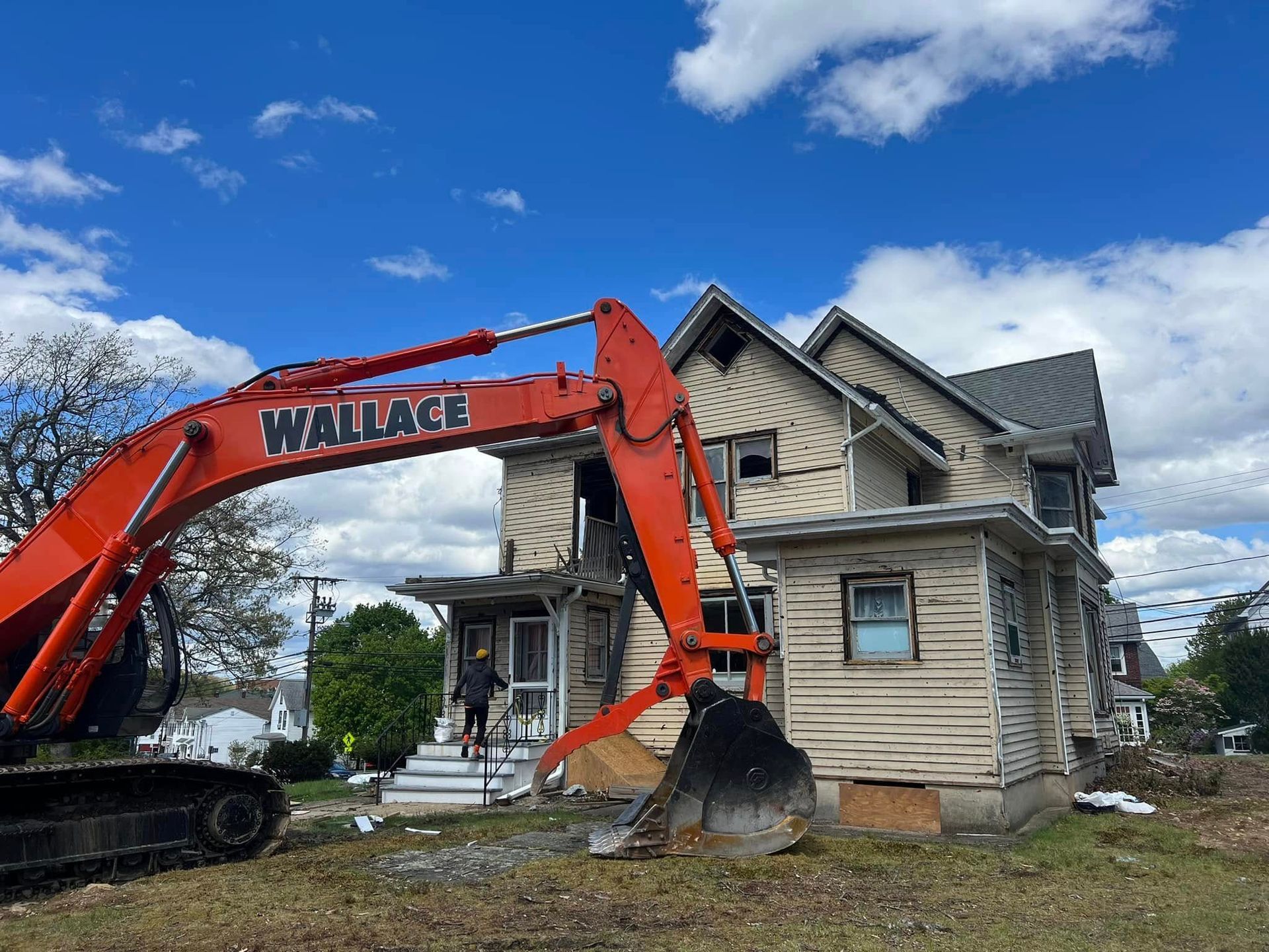 Orange excavator demolishing a two-story beige house under a blue sky. Person on the steps.