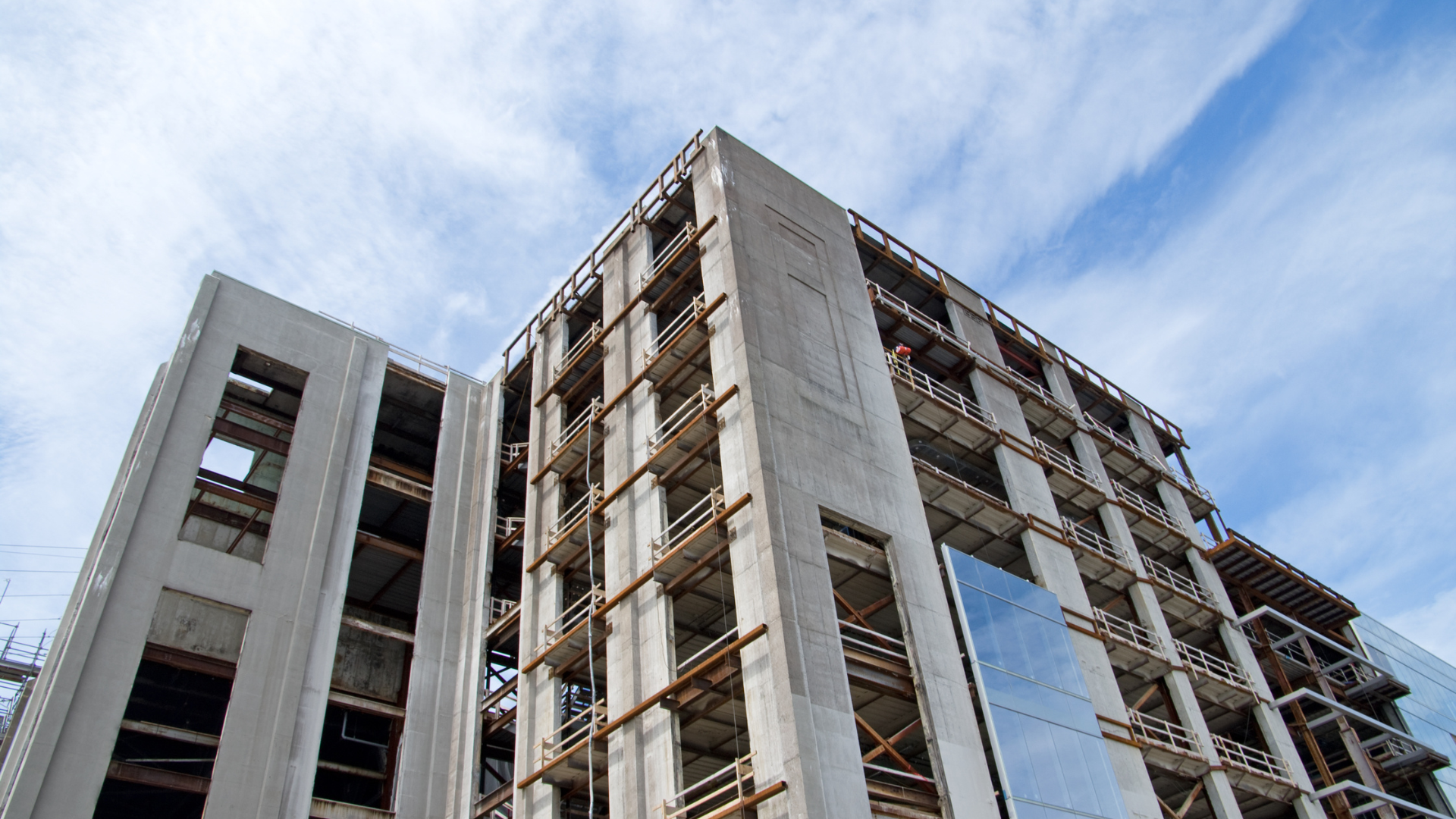 Building under construction with concrete walls, scaffolding, and glass, set against a cloudy blue sky.