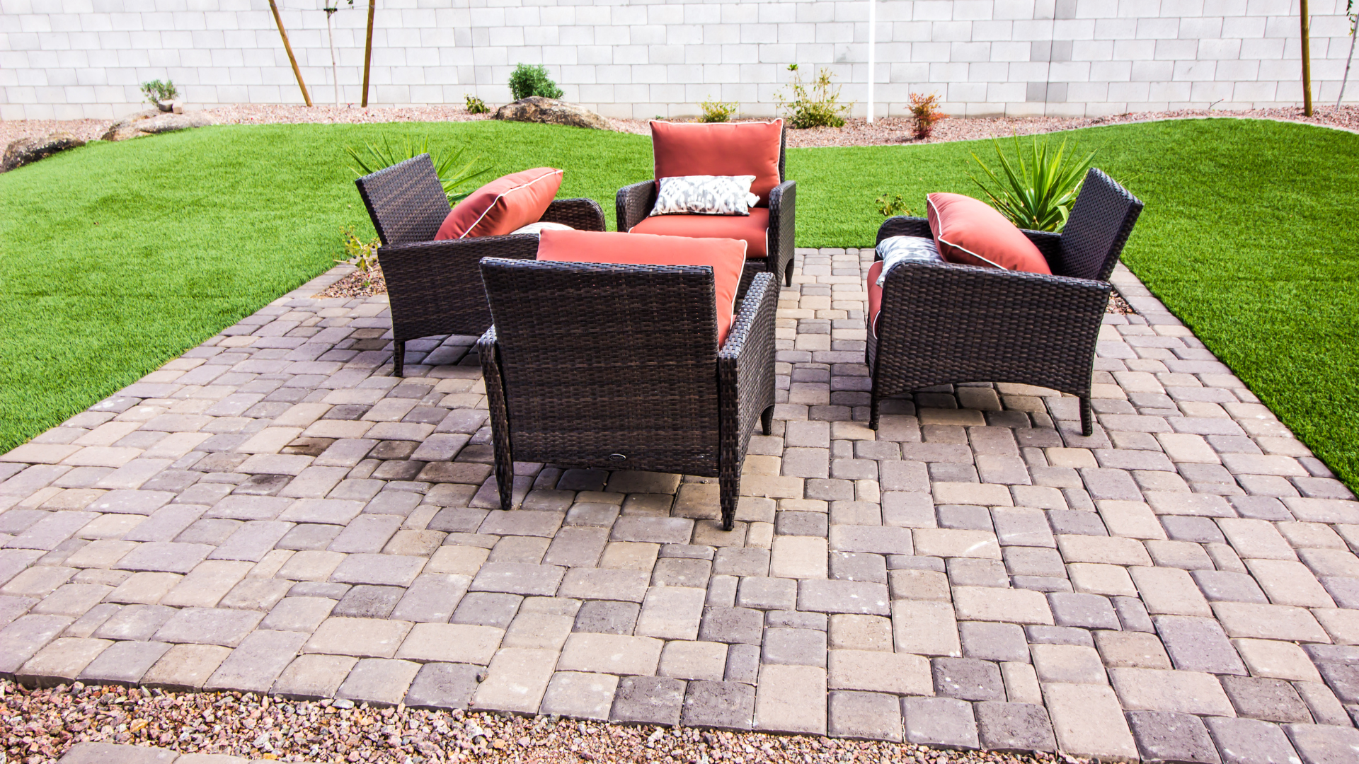 Patio seating area with brick pavers, wicker chairs, and orange cushions, near a lawn.