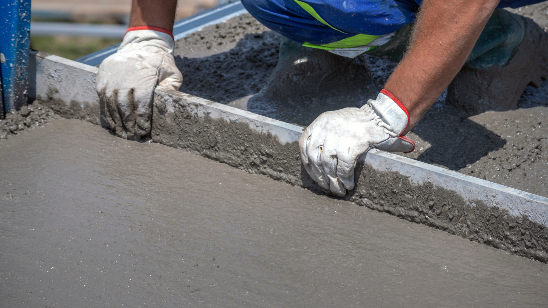 A worker in gloves screeds wet concrete with a long tool, leveling the surface.