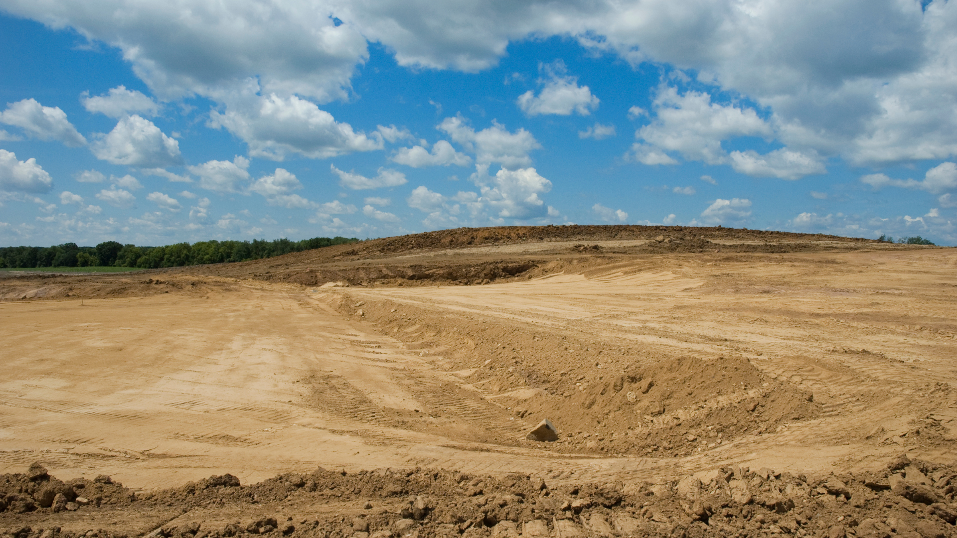 A large, barren plot of land with brown dirt, under a blue sky with white clouds.