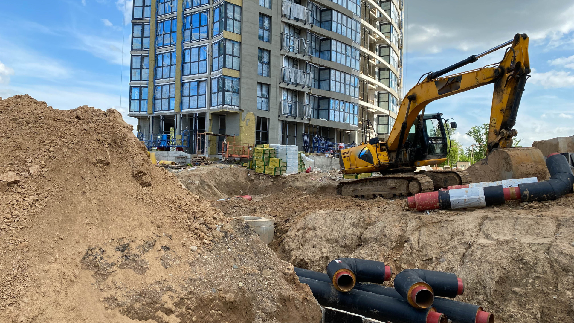 Construction site with excavator and unfinished building; dirt and pipes in foreground.